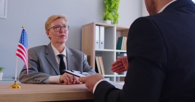 Male person sitting in the office of the US public services or embassy having consular visa interview with mature woman officer with US flag at the desk of official's workplace. 4k video.