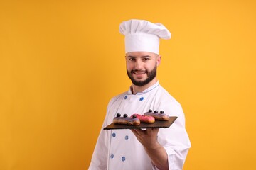 Happy confectioner in uniform holding eclairs on orange background
