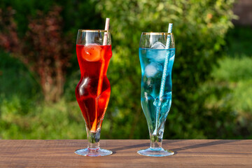 Naklejka na ściany i meble Two artistic glasses shaped like a nude woman and man filled with red and blue alcoholic cocktails on table with lush green garden background, closeup. Playful summer vibe for creative drink concepts