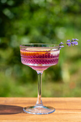 Bright cocktail or lemonade served in elegant crystal glass on wooden table against of a green summer garden. Purple color drink with lavender herb flowers and slice of lemon, closeup