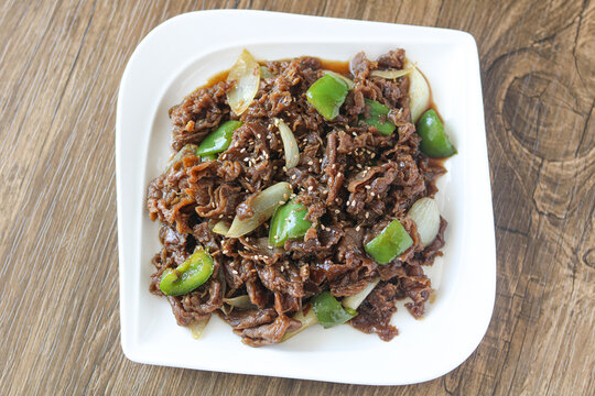 Marinated beef slices stir-fried with green bell peppers and onions, sprinkled with white sesame seeds.&nbsp;The dish is presented on a white rectangular plate set on a wooden background.