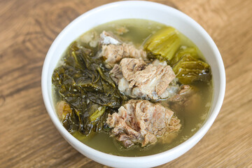 Close-up of a hearty bowl of Bak Kut served on a rustic wooden background. The savory broth contains tender pork ribs and large pieces of green leafy vegetables.