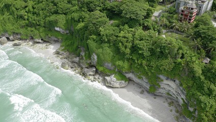 Aerial view of a secluded beach with a rocky shoreline, lush greenery, and a modern building nestled in the cliffside