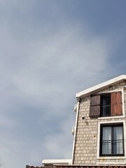 Stone house with open wooden shutters and white roof, photographed from below against blue sky with white clouds. Buljarica, Montenegro – July 26, 2025. Editorial use only.
