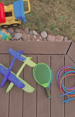 Backyard photo with a pile of outdoor toys for kids. A bunch of sports toys are lying on a wooden patio. Tennis rackets, plastic hoops, planes made of foam, and toy vehicle are beloved toys for boys. 