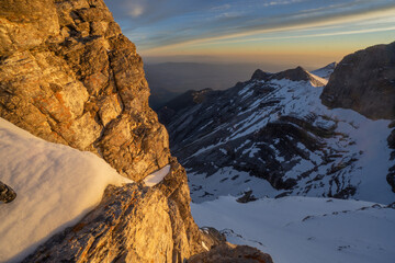 Aerial view of jagged peaks dusted with fresh snow catch the warm glow of the setting sun, contrasting with the cool shadows in the valleys below, location data omitted.