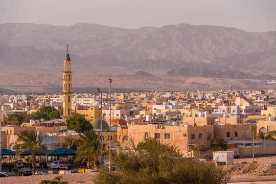 Panorama of Aqaba, a large Jordanian port city on the Red Sea, with a mosque minaret and colorful buildings set against the Israeli mountains at sunset.
