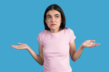 Puzzled young woman shrugging shoulders and looking up on blue background