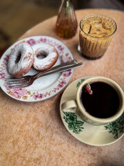 donuts dusted with powdered sugar on a floral plate, served with iced coffee and hot black coffee on a vintage-style café table.