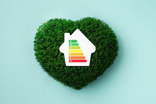 A house model with energy efficiency scale on green moss heart and light blue background. Top view, flat lay.