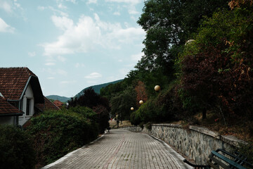 Stone walkway in Mokra Gora, Serbia, lined with bushes, trees and round street lamps.