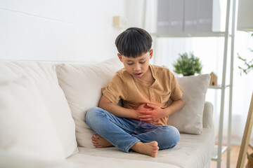 Fototapeta premium Portrait of sick unhappy little child asian boy touching belly stomach with suffering from stomach ache pain, gastritis, gastroenteritis, food poisoning, diarrhea, intestinal inflammation at home