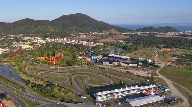 Wide aerial view of Beto Carrero World in Penha, Brazil, showing the winding go-kart racing track, colorful roller coaster, and drop tower framed by lush green hills and coastal surroundings.