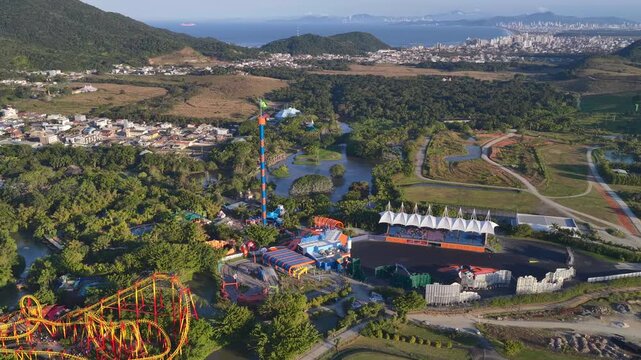 Drone view above Beto Carrero World in Penha, Brazil, showing colorful roller coasters, tall drop tower, water canals, and themed architecture amid green surroundings and distant urban skyline.