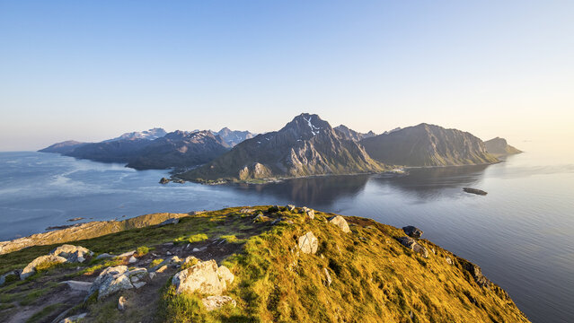 View of rugged, snow-capped mountains meeting the serene, reflective waters from the grassy foreground, bathed in the soft glow of dawn, Leknes, Nordland, Norway.