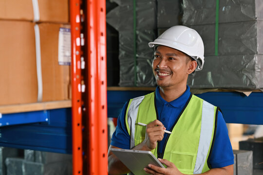 Warehouse employee in safety gear checking inventory on shelves with a pen and tablet, smiling while managing stock