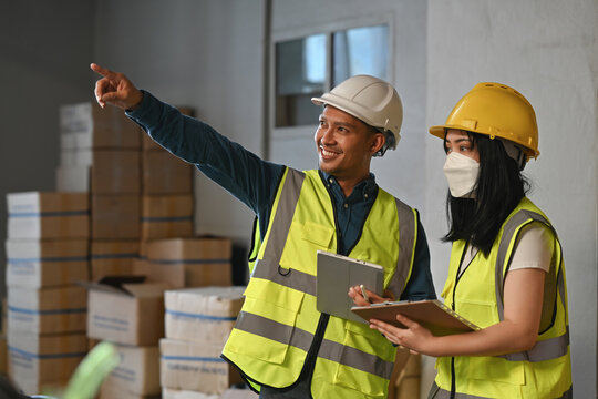 Two warehouse workers in safety vests and helmets planning operations with tablet and clipboard inside storage facility