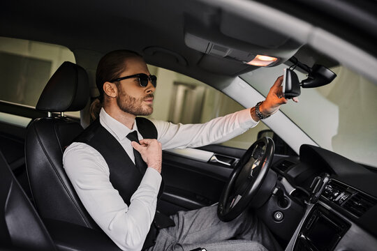 Handsome man in elegant suit sits in luxury car, adjusting mirror while parked underground.