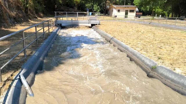  Canal de esgoto de uma esta&ccedil;&atilde;o de tratamento de &aacute;gua, mostrando &aacute;gua turva a fluir atrav&eacute;s de uma pequena barragem com um rastelo