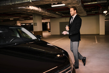 A handsome man in elegant attire approaches his luxury car with a cup of coffee, exuding confidence.