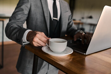 A man in a suit holds a coffee cup while working on his laptop in a stylish cafe.