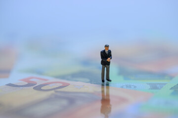 Miniaturgeschäftsmann steht auf unscharfen Euroscheinen – Symbolbild für finanzielle Unsicherheit und Machtverhältnisse