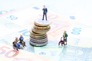 Miniaturgeschäftsmann auf Münzstapel hinter Rentnern auf Parkbank – Symbolbild soziale Ungleichheit