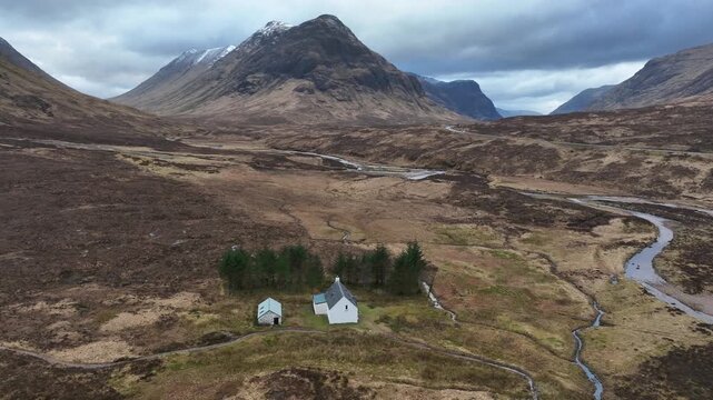 Aerial view of a white house and outbuilding near Buachaille Etive M&ograve;r, nestled in a valley with a winding stream under a cloudy sky, Ballachulish, Scotland, United Kingdom.