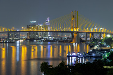 Fototapeta premium Scenery view after sunset of Thotsamarachan Bridge (Rama IX Bridge) crossing Chao phraya river with skyscraper in the background. Beautiful city of Bangkok with bright glowing lights in the sundown.