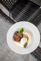 Grilled meat patty with mashed potatoes, poached egg, and greens on a white plate placed on a wicker chair, presented in a rustic dining setting.
