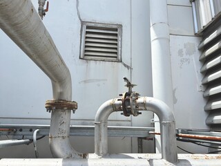 An industrial scene with a complex network of pipes and valves against a weathered white wall. The metallic surfaces and vent show the robust, functional design of a factory or mechanical system.