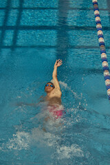 A swimmer performs a backstroke in an indoor pool adjacent to a lane divider, demonstrating...