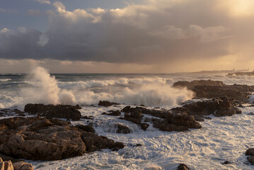 Rough seas crash against rocky shores along the Whale Coast, Hermanus, in the scenic Overberg region of Western Cape, South Africa, under dramatic cloudy skies and vibrant coastal vegetation.