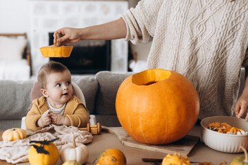 Happy Halloween. Happy mother carving halloween pumpkin together with baby son on table with spooky halloween decor. Family making jack o lantern, holiday joyful time