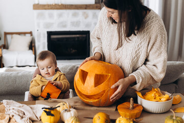 Family carving halloween pumpkin. Happy mother and cute baby son cutting pumpkin on table with ghosts, spiders and bats decoration. Making a jack o lantern, halloween preparation