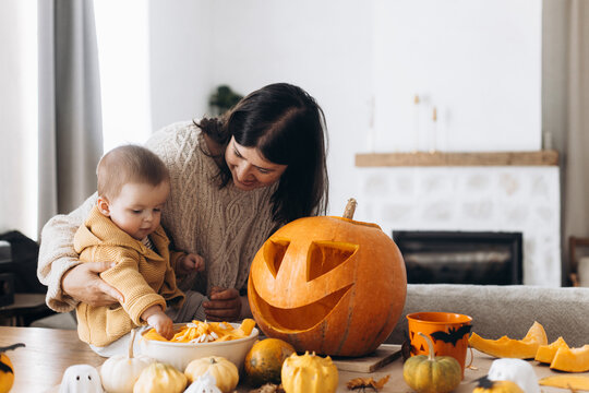 Family carving halloween pumpkin. Happy mother and cute baby son cutting pumpkin on table with ghosts, spiders and bats decoration. Making a jack o lantern, halloween preparation