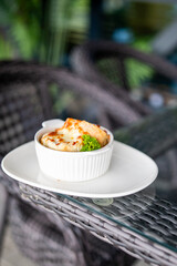 Baked dish served in a white ceramic bowl with parsley garnish, placed on a plate over glass table in outdoor setting with wicker furniture and blurred greenery.