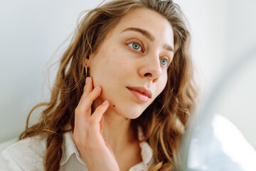 Fototapeta premium Portrait of a young woman looking at a small mirror in a cozy room. Beautiful woman touches her face with her hands and enjoys natural beauty. Concept of beauty, dermatology, comfort.