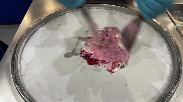 Street vendor preparing fresh rolled ice cream on a cold pan using metal spatulas, mixing creamy base with chocolate, red berry sauce and toppings