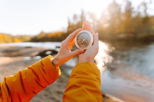 Close-up of young female hiker's hands holding compass against lake background. Woman in bright jacket enjoying sunset and looking for hiking direction on sunny autumn trail. Navigation concept. - Powered by Adobe