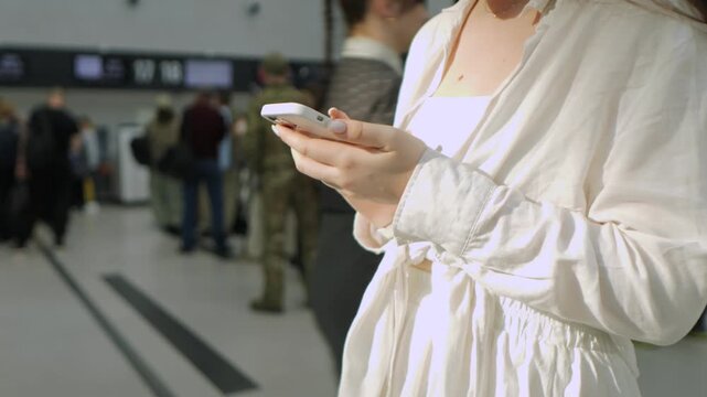 Woman is holding a cell phone in a busy airport. She is wearing a white jacket and she is focused on her phone. The scene is bustling with people and luggage, creating a sense of urgency