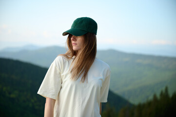 Woman walks on grassy mountain trail under warm sun, with wooden cabins and distant hills in...