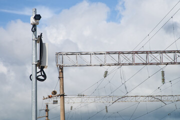 A modern white surveillance video camera on a metal pole beside electrified railway tracks, framed by overhead power cables under a cloudy sky. Photo