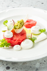 Artfully plated gourmet appetizer with watermelon, avocado, cream, and herbs on a white dish. Vibrant colors and elegant styling ideal for culinary themes.