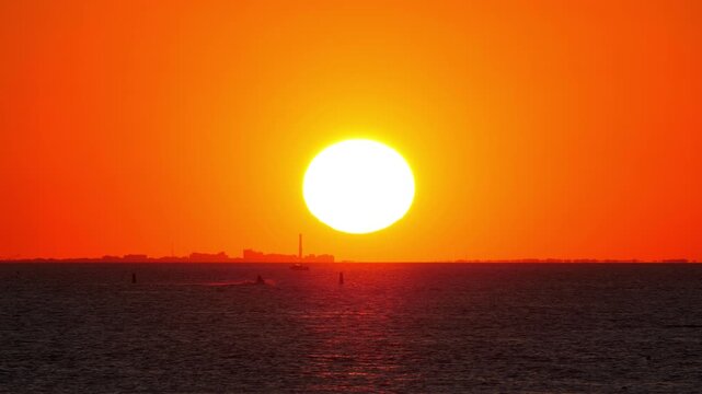 Large sun disk sets over bay in telephoto shot, casting orange glow over calm water. In distance, jet ski moves almost in silhouette, adding some motion to this serene and picturesque scene