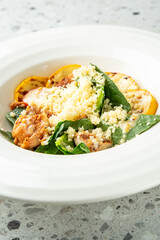 Colorful healthy meal with grilled squash, chicken, couscous, spinach, and cherry tomatoes served on a white plate over a speckled countertop, top view.