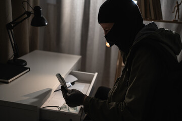 Illegally breaking. Male robber wearing black balaclava steals money from desk at apartment. Thief...