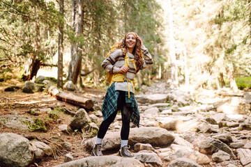 Naklejka premium Female hiker with a colorful hiking backpack on a walking rocky trail in the mountains. Cute woman enjoying mountain scenery in a dense forest. Adventure, hiking and nature concept.