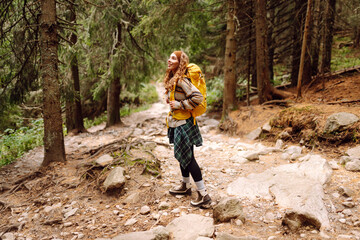 Obraz premium Female hiker with a colorful hiking backpack on a walking rocky trail in the mountains. Cute woman enjoying mountain scenery in a dense forest. Adventure, hiking and nature concept.
