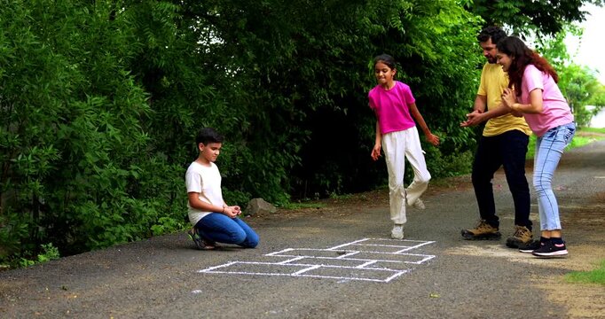 Indian family playing Tikkar Billa, a traditional hopscotch game in home garden, spending quality fun time together with children hopping on squares and enjoying nostalgic outdoor sports activity